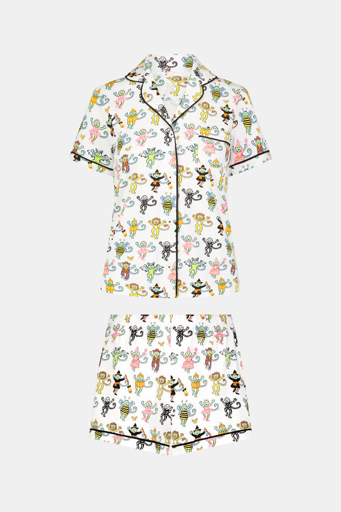 A smiling child in Roller Rabbit’s Kids Boo Crew Pajamas, crafted from soft Pima cotton, sits with arms raised on outdoor steps among women in coordinating patterned pajamas, all sharing a cheerful and relaxed moment together.