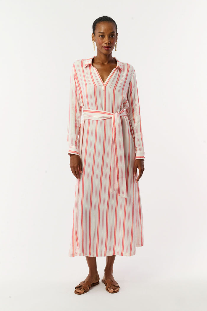 A woman with short hair and gold earrings wears the Roller Rabbit Cabana Stripe Fallon Dress—an ankle-length, long-sleeve vacation dress with vertical red stripes and a matching waist tie—paired with brown sandals against a white background.