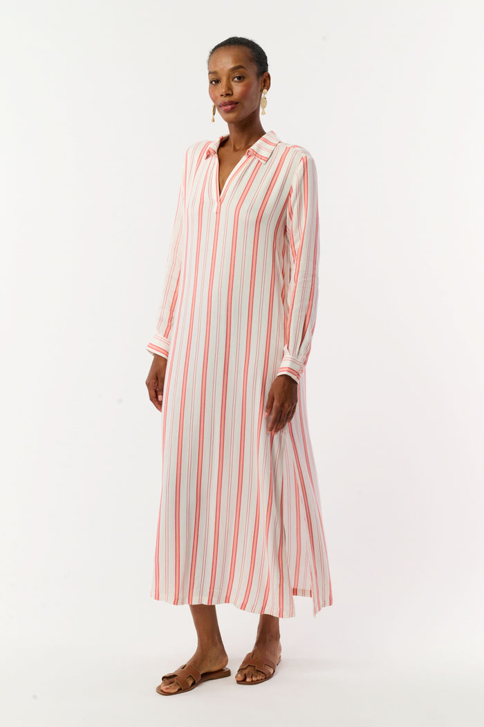 A woman with short hair and gold earrings wears the Roller Rabbit Cabana Stripe Fallon Dress—an ankle-length, long-sleeve vacation dress with vertical red stripes and a matching waist tie—paired with brown sandals against a white background.