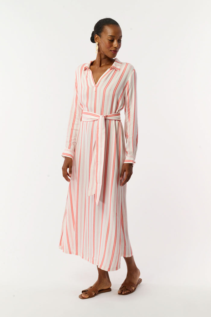 A woman with short hair and gold earrings wears the Roller Rabbit Cabana Stripe Fallon Dress—an ankle-length, long-sleeve vacation dress with vertical red stripes and a matching waist tie—paired with brown sandals against a white background.