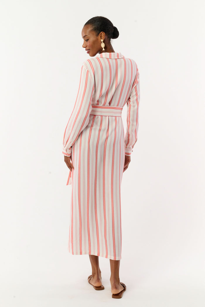 A woman with short hair and gold earrings wears the Roller Rabbit Cabana Stripe Fallon Dress—an ankle-length, long-sleeve vacation dress with vertical red stripes and a matching waist tie—paired with brown sandals against a white background.
