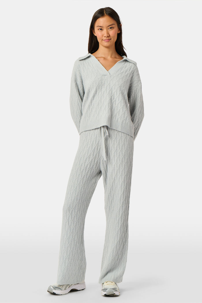 A woman wearing a light gray Cable Knots Rocio Sweater by Roller Rabbit, featuring a textured knit and V-neck, paired with matching knit pants, stands against a plain white background.