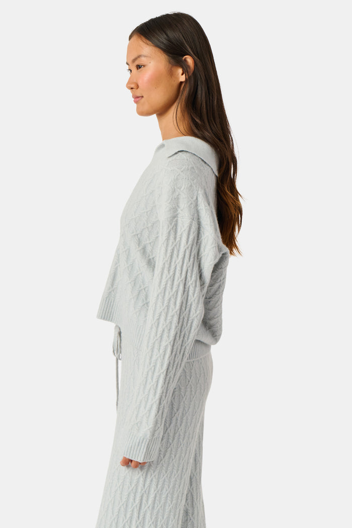 A woman wearing a light gray Cable Knots Rocio Sweater by Roller Rabbit, featuring a textured knit and V-neck, paired with matching knit pants, stands against a plain white background.