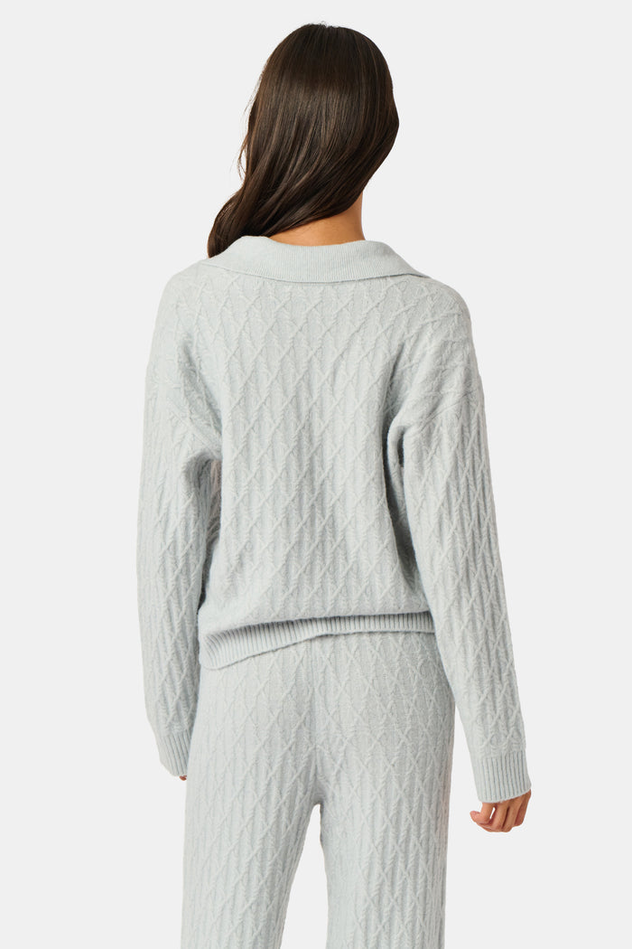 A woman wearing a light gray Cable Knots Rocio Sweater by Roller Rabbit, featuring a textured knit and V-neck, paired with matching knit pants, stands against a plain white background.