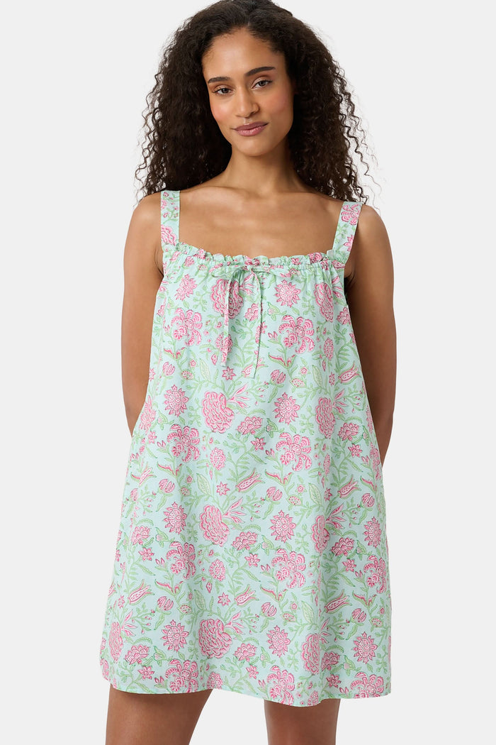 A woman with curly hair wears the Camellia Block Keaton Dress by Roller Rabbit, a sleeveless, loose-fit 100 cotton sundress with pink floral print on a light green background. She smiles softly with her hands behind her back.