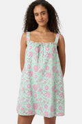 A woman with curly hair wears the Camellia Block Keaton Dress by Roller Rabbit, a sleeveless, loose-fit 100 cotton sundress with pink floral print on a light green background. She smiles softly with her hands behind her back.