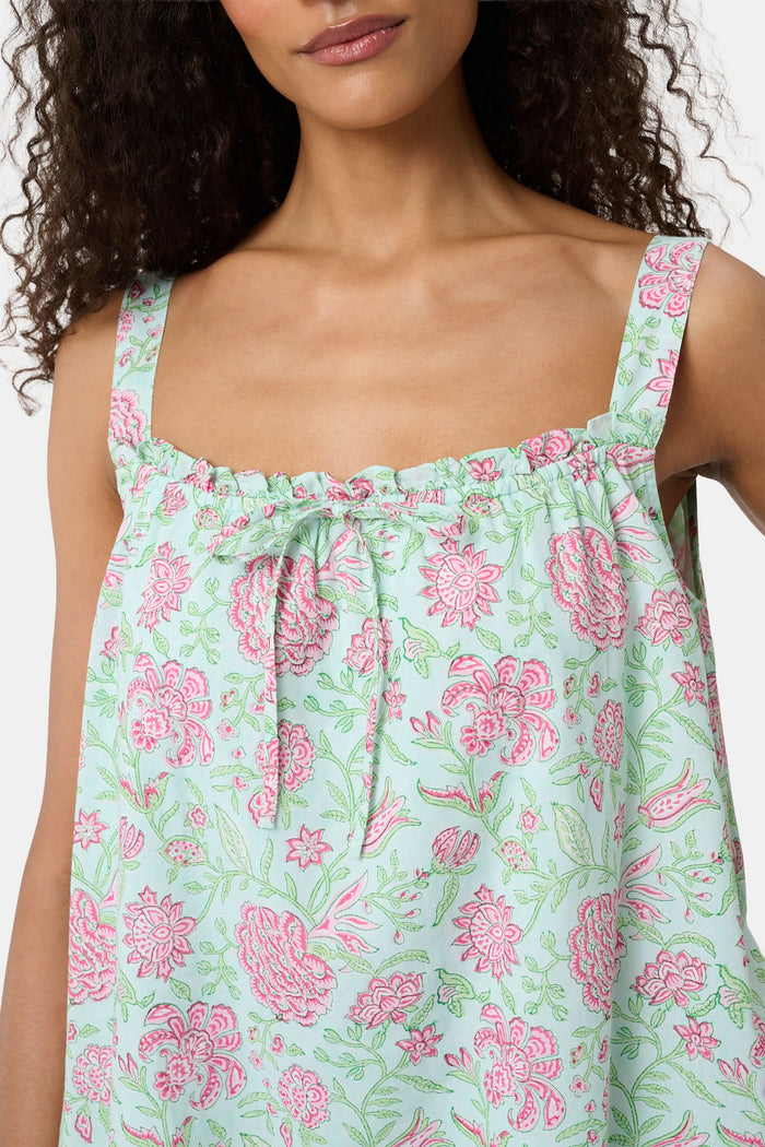 A woman with curly hair wears the Camellia Block Keaton Dress by Roller Rabbit, a sleeveless, loose-fit 100 cotton sundress with pink floral print on a light green background. She smiles softly with her hands behind her back.