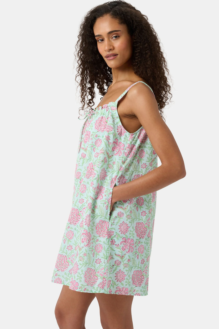 A woman with curly hair wears the Camellia Block Keaton Dress by Roller Rabbit, a sleeveless, loose-fit 100 cotton sundress with pink floral print on a light green background. She smiles softly with her hands behind her back.