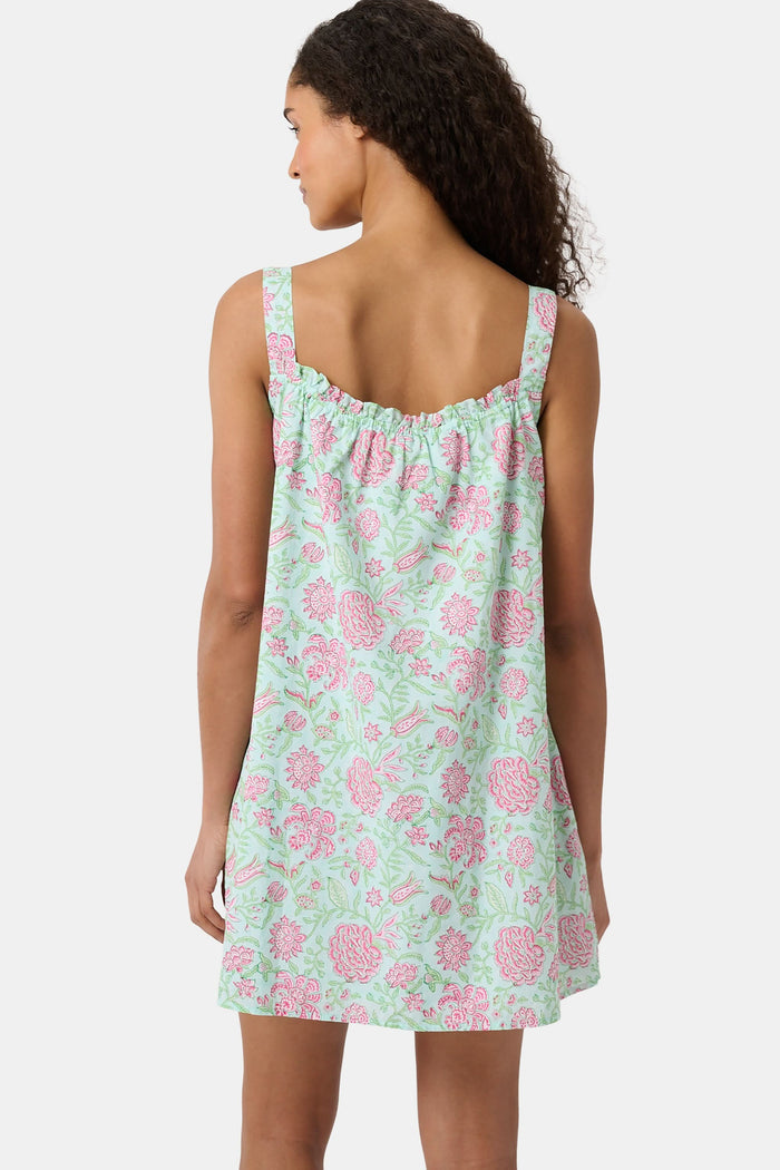 A woman with curly hair wears the Camellia Block Keaton Dress by Roller Rabbit, a sleeveless, loose-fit 100 cotton sundress with pink floral print on a light green background. She smiles softly with her hands behind her back.