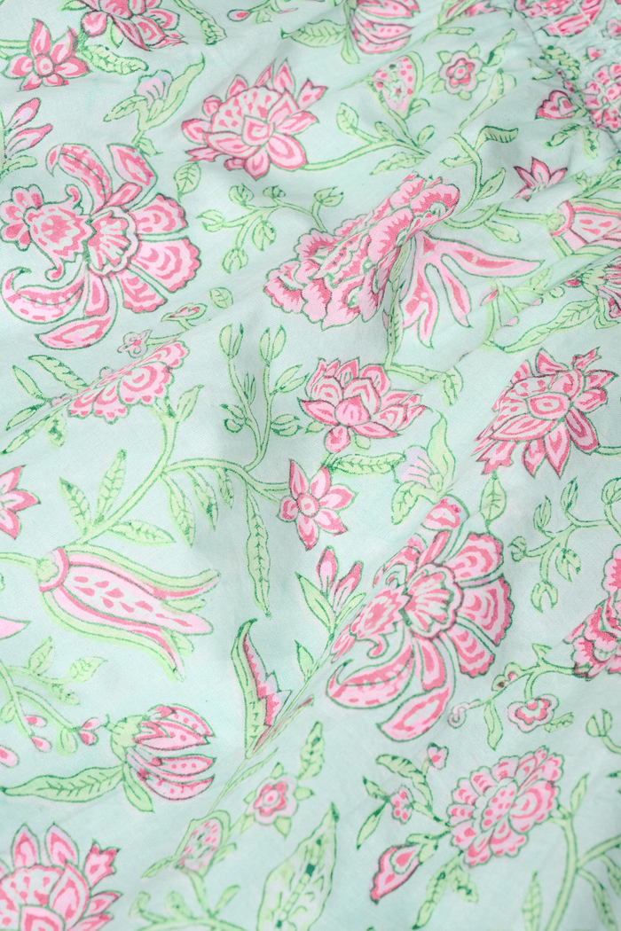 A woman with curly hair wears the Camellia Block Keaton Dress by Roller Rabbit, a sleeveless, loose-fit 100 cotton sundress with pink floral print on a light green background. She smiles softly with her hands behind her back.