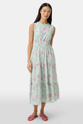 A woman with long dark hair and a neutral expression poses against a plain light background wearing the Roller Rabbit Camellia Block Milani Dress, a pastel green and pink floral cotton maxi, paired with pink sandals.