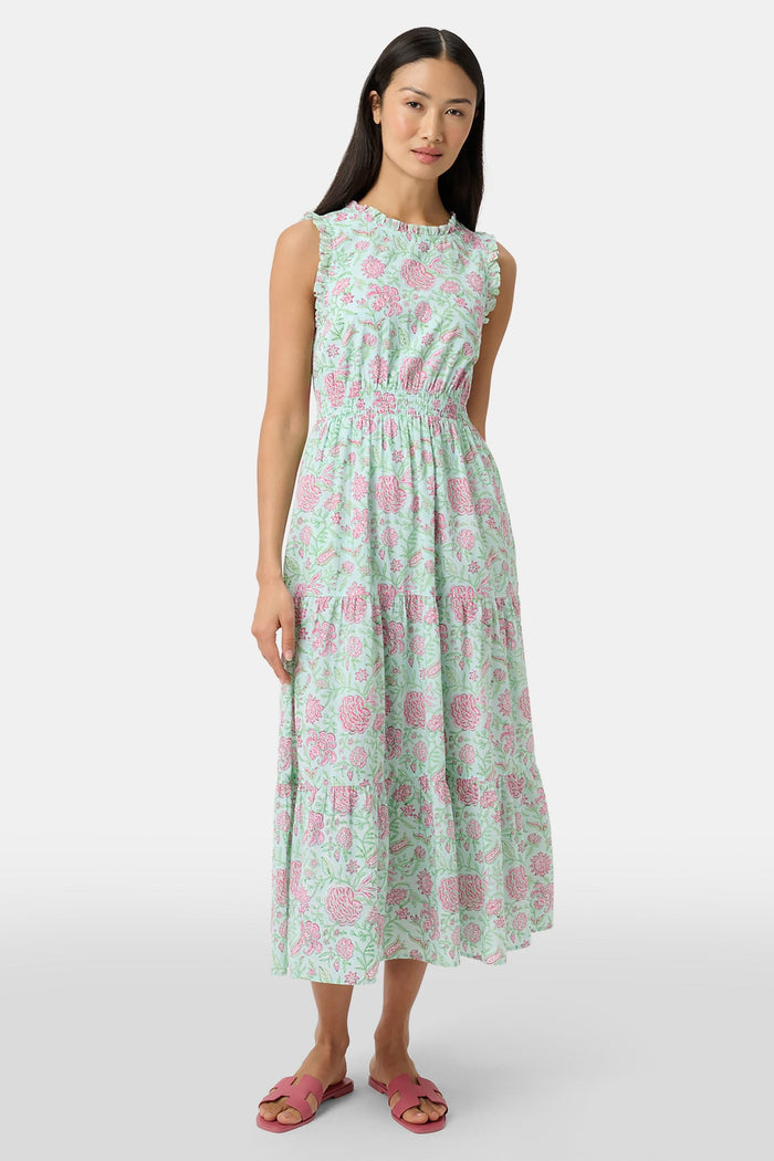 A woman with long dark hair and a neutral expression poses against a plain light background wearing the Roller Rabbit Camellia Block Milani Dress, a pastel green and pink floral cotton maxi, paired with pink sandals.