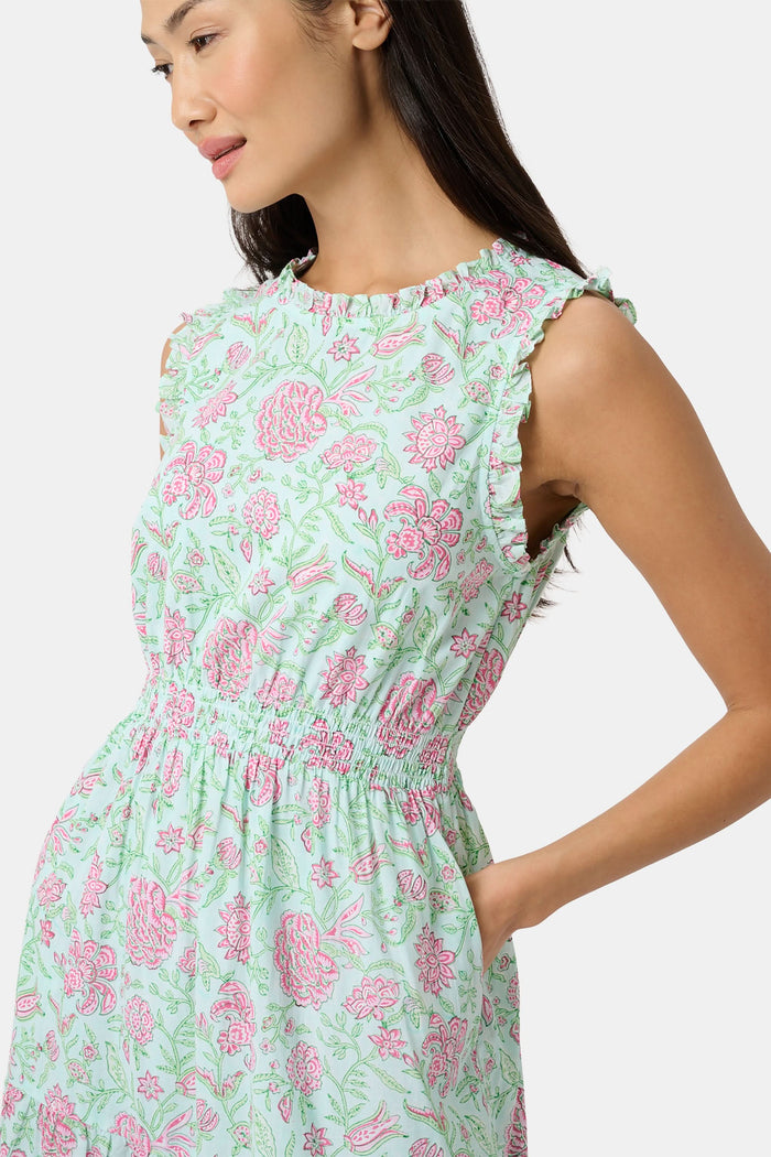 A woman with long dark hair and a neutral expression poses against a plain light background wearing the Roller Rabbit Camellia Block Milani Dress, a pastel green and pink floral cotton maxi, paired with pink sandals.