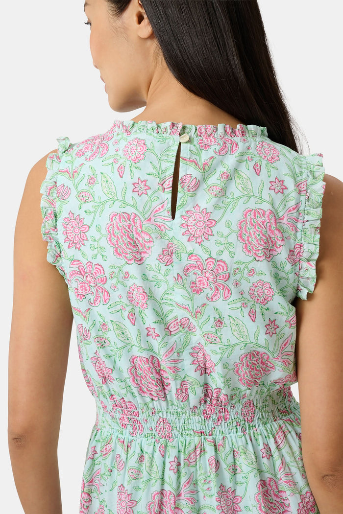 A woman with long dark hair and a neutral expression poses against a plain light background wearing the Roller Rabbit Camellia Block Milani Dress, a pastel green and pink floral cotton maxi, paired with pink sandals.