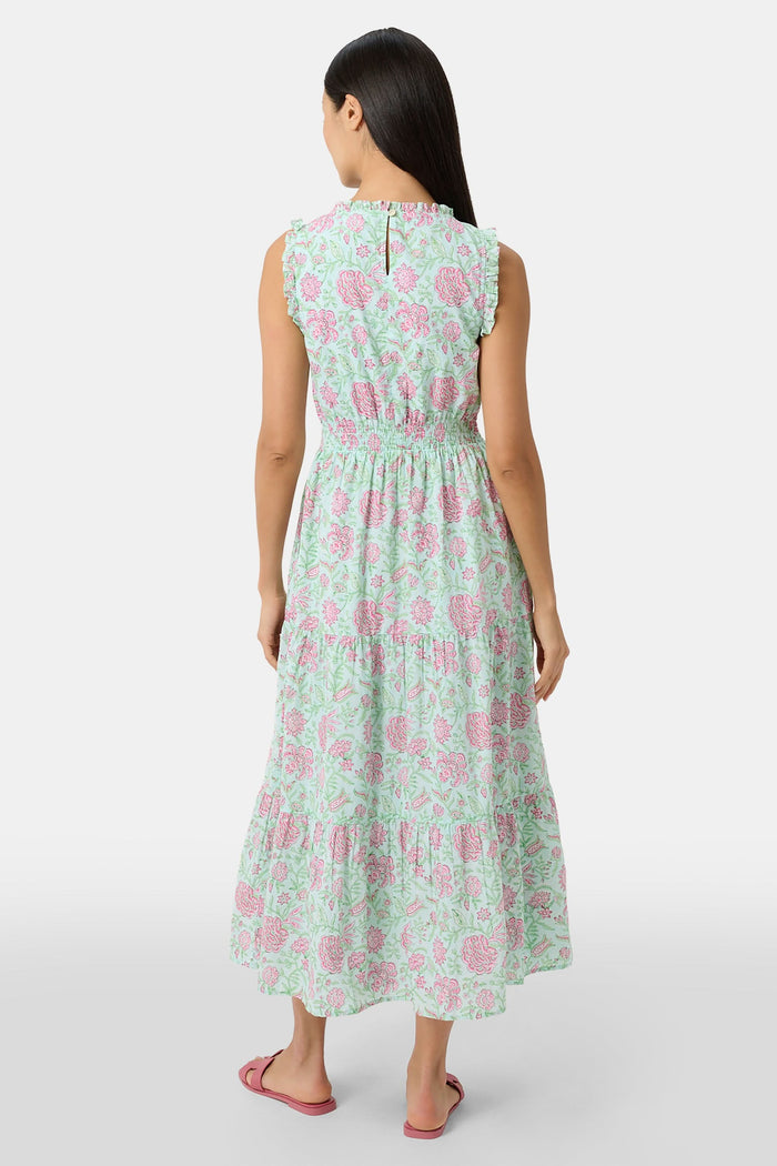 A woman with long dark hair and a neutral expression poses against a plain light background wearing the Roller Rabbit Camellia Block Milani Dress, a pastel green and pink floral cotton maxi, paired with pink sandals.