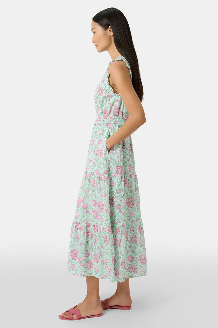 A woman with long dark hair and a neutral expression poses against a plain light background wearing the Roller Rabbit Camellia Block Milani Dress, a pastel green and pink floral cotton maxi, paired with pink sandals.