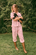 Wearing the Charlie Liza Set by Roller Rabbit, a woman stands on the grass in brown boots, holding a small black animal, with green foliage in the background.