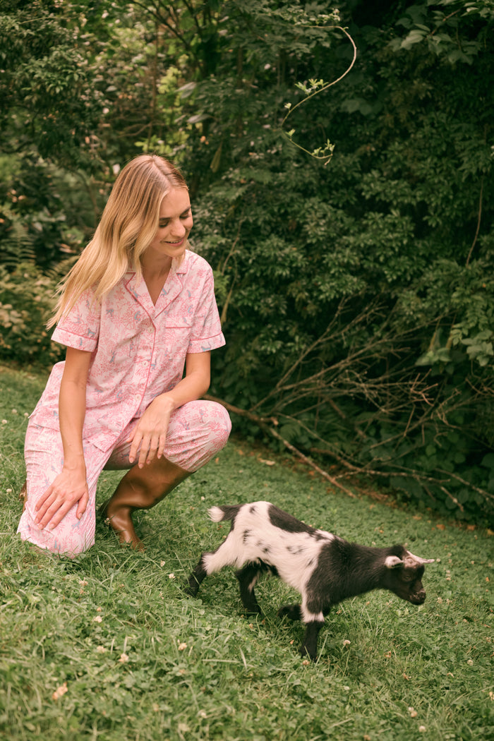 Wearing the Charlie Liza Set by Roller Rabbit, a woman stands on the grass in brown boots, holding a small black animal, with green foliage in the background.
