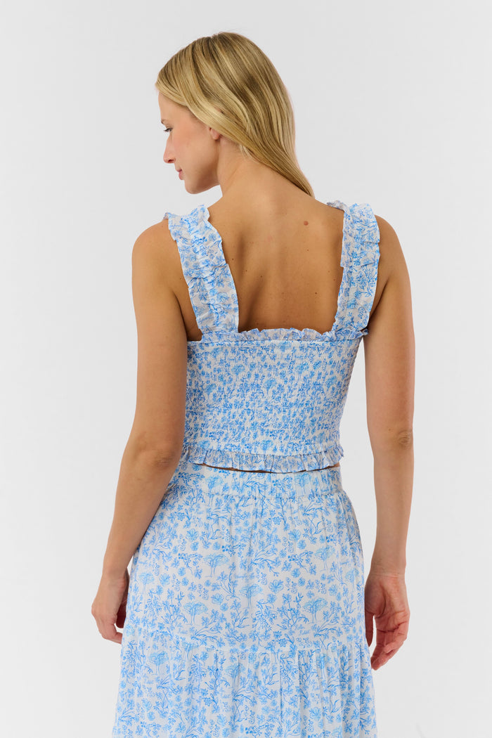 A woman wears the Roller Rabbit Cherry Blossom Toile Sabrina Top—a light blue and white floral smocked crop with ruffled straps and a square neckline. Only her upper body is shown against a plain background.