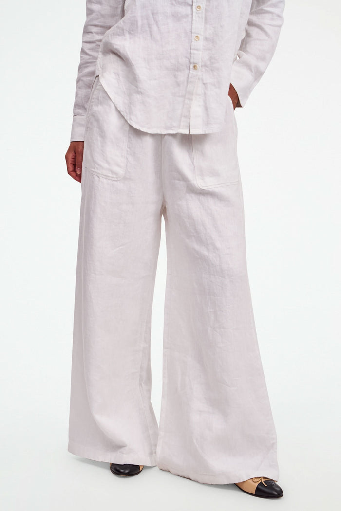 A person wearing Roller Rabbit's Chester Pant—loose-fitting linen wide leg pants with an elastic waist—paired with a matching white button-up shirt and two-tone black and beige flats, shown from shoulders to feet with one hand in their pocket.