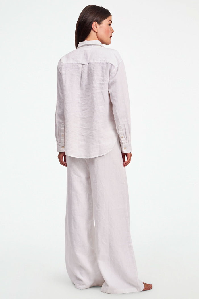 A person wearing Roller Rabbit's Chester Pant—loose-fitting linen wide leg pants with an elastic waist—paired with a matching white button-up shirt and two-tone black and beige flats, shown from shoulders to feet with one hand in their pocket.