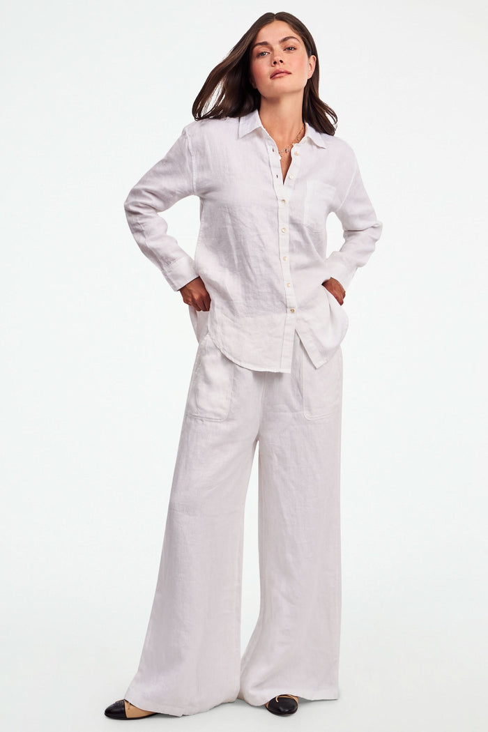 A person wearing Roller Rabbit's Chester Pant—loose-fitting linen wide leg pants with an elastic waist—paired with a matching white button-up shirt and two-tone black and beige flats, shown from shoulders to feet with one hand in their pocket.
