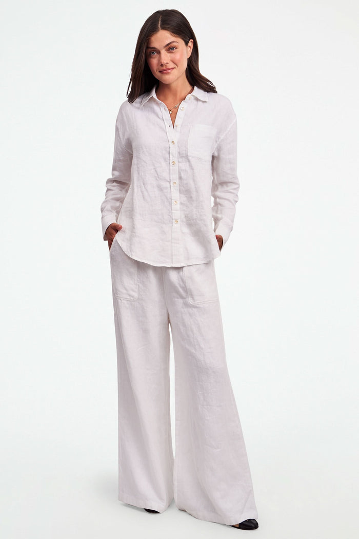 A person wearing Roller Rabbit's Chester Pant—loose-fitting linen wide leg pants with an elastic waist—paired with a matching white button-up shirt and two-tone black and beige flats, shown from shoulders to feet with one hand in their pocket.