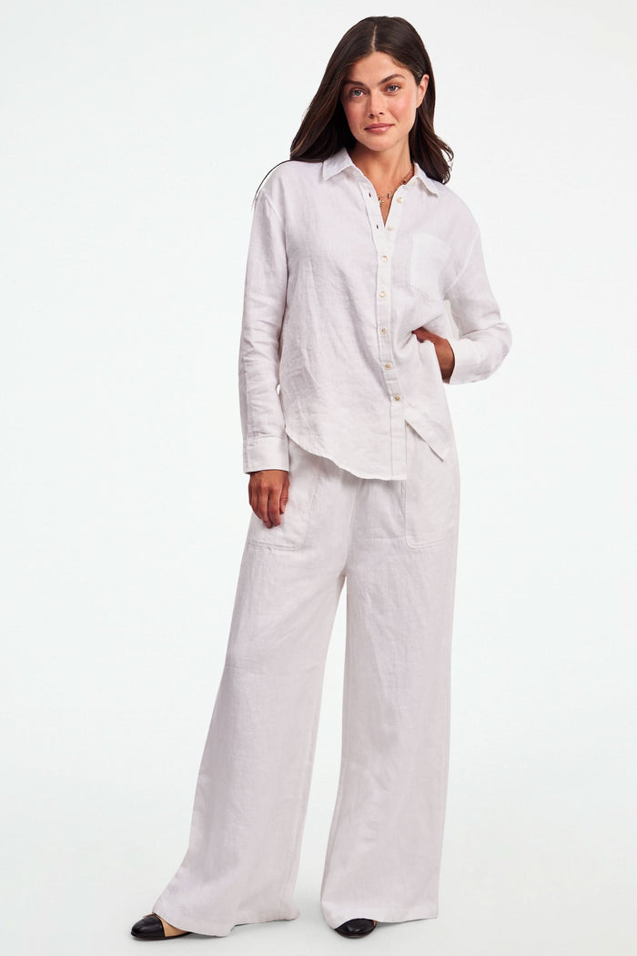 A person wearing Roller Rabbit's Chester Pant—loose-fitting linen wide leg pants with an elastic waist—paired with a matching white button-up shirt and two-tone black and beige flats, shown from shoulders to feet with one hand in their pocket.