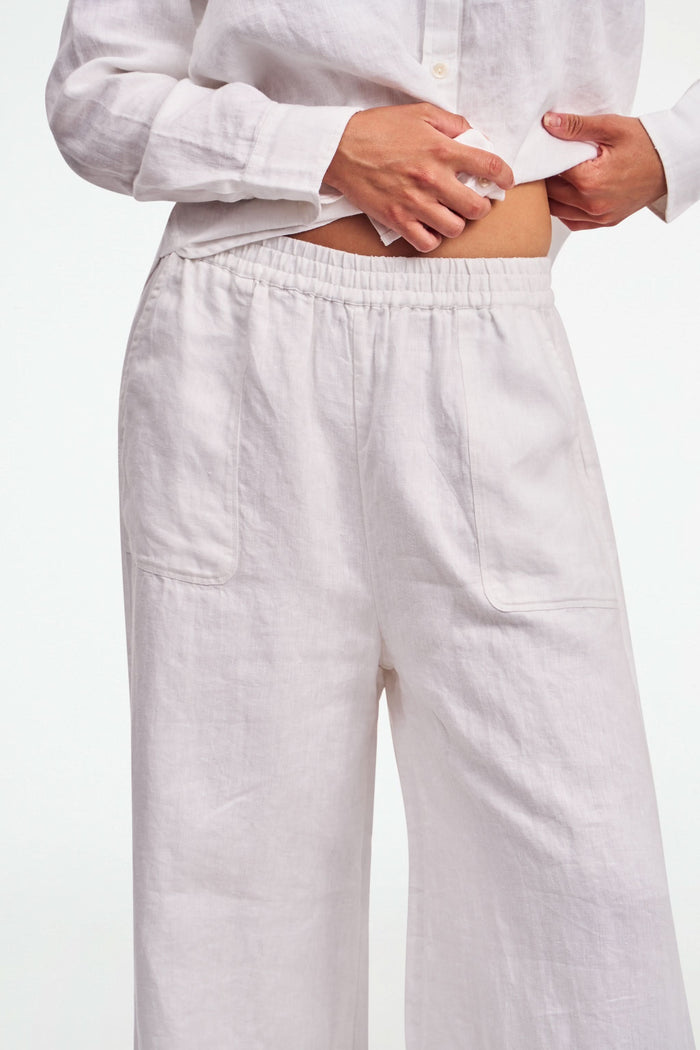 A person wearing Roller Rabbit's Chester Pant—loose-fitting linen wide leg pants with an elastic waist—paired with a matching white button-up shirt and two-tone black and beige flats, shown from shoulders to feet with one hand in their pocket.