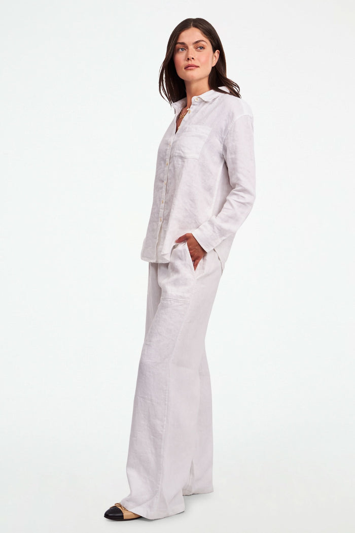 A person wearing Roller Rabbit's Chester Pant—loose-fitting linen wide leg pants with an elastic waist—paired with a matching white button-up shirt and two-tone black and beige flats, shown from shoulders to feet with one hand in their pocket.