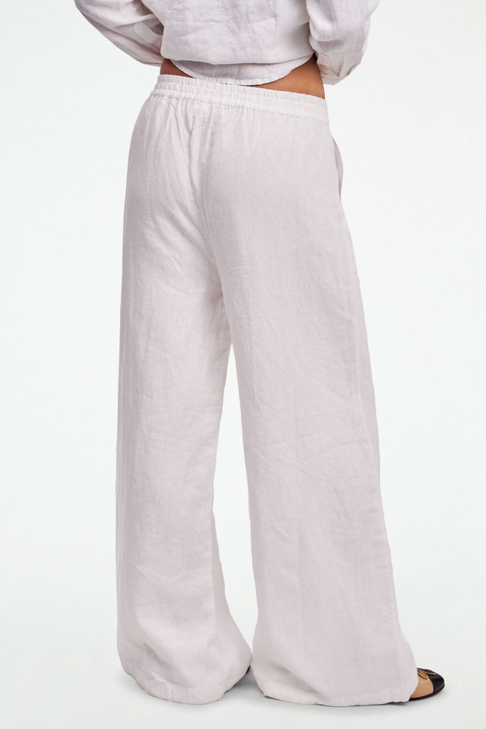 A person wearing Roller Rabbit's Chester Pant—loose-fitting linen wide leg pants with an elastic waist—paired with a matching white button-up shirt and two-tone black and beige flats, shown from shoulders to feet with one hand in their pocket.