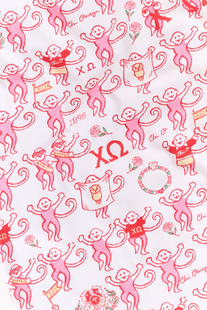 A young woman with red hair sits on a bench outside, holding a phone and earphones while looking to the side. She is wearing the Roller Rabbit Chi Omega Monkey Polo Pajamas, which feature a front button placket and an elastic waistband in a pink hue. A light jacket is draped over the bench beside her.