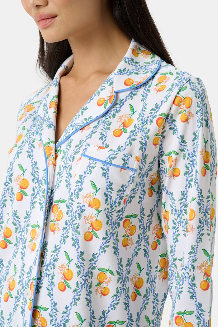A woman wearing Roller Rabbit's Clementine Trellis Long Sleeve Polo Pajamas stands in a garden with a bouquet, beside blooming pink flowers and a wicker basket filled with oranges and peaches on the grass.