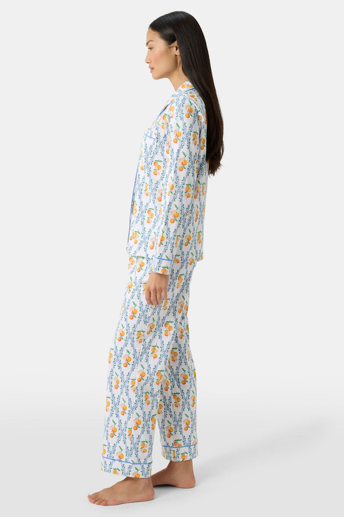 A woman wearing Roller Rabbit's Clementine Trellis Long Sleeve Polo Pajamas stands in a garden with a bouquet, beside blooming pink flowers and a wicker basket filled with oranges and peaches on the grass.