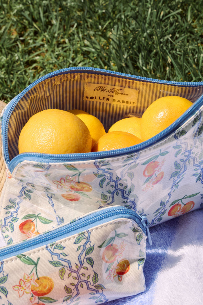 The Clementine Trellis Makeup Bag by Roller Rabbit is a zippered, water-resistant cotton canvas bag with a blue zipper and a white background featuring blue geometric lines, green leaves, orange fruits, and white flowers.