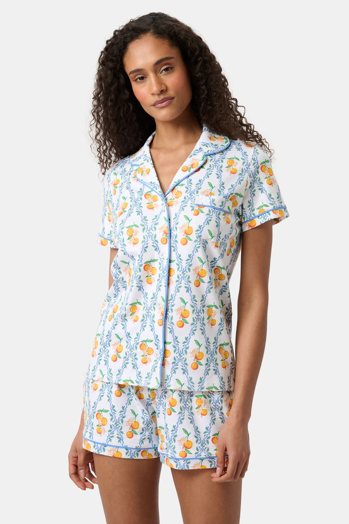 A woman with long hair strolls on the grass in a garden, wearing the Clementine Trellis Polo Pajamas by Roller Rabbit. Green plants and purple flowers bloom in the sunlight behind her.