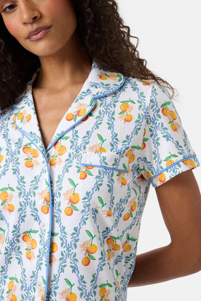 A woman with long hair strolls on the grass in a garden, wearing the Clementine Trellis Polo Pajamas by Roller Rabbit. Green plants and purple flowers bloom in the sunlight behind her.