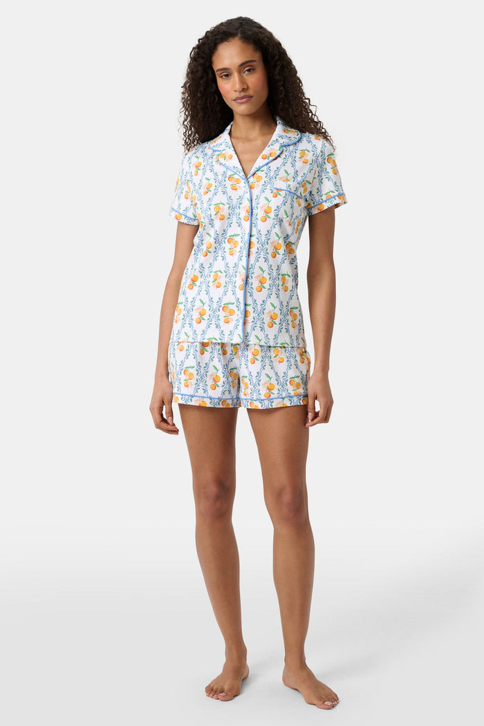 A woman with long hair strolls on the grass in a garden, wearing the Clementine Trellis Polo Pajamas by Roller Rabbit. Green plants and purple flowers bloom in the sunlight behind her.