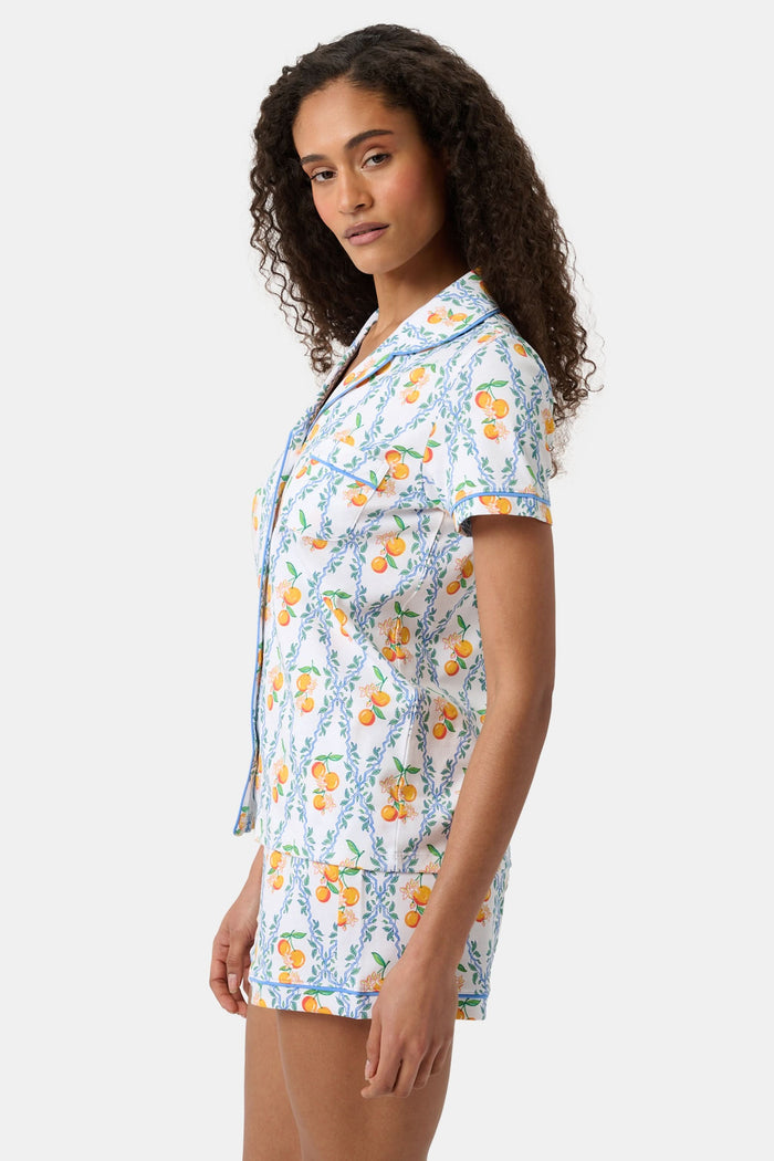 A woman with long hair strolls on the grass in a garden, wearing the Clementine Trellis Polo Pajamas by Roller Rabbit. Green plants and purple flowers bloom in the sunlight behind her.