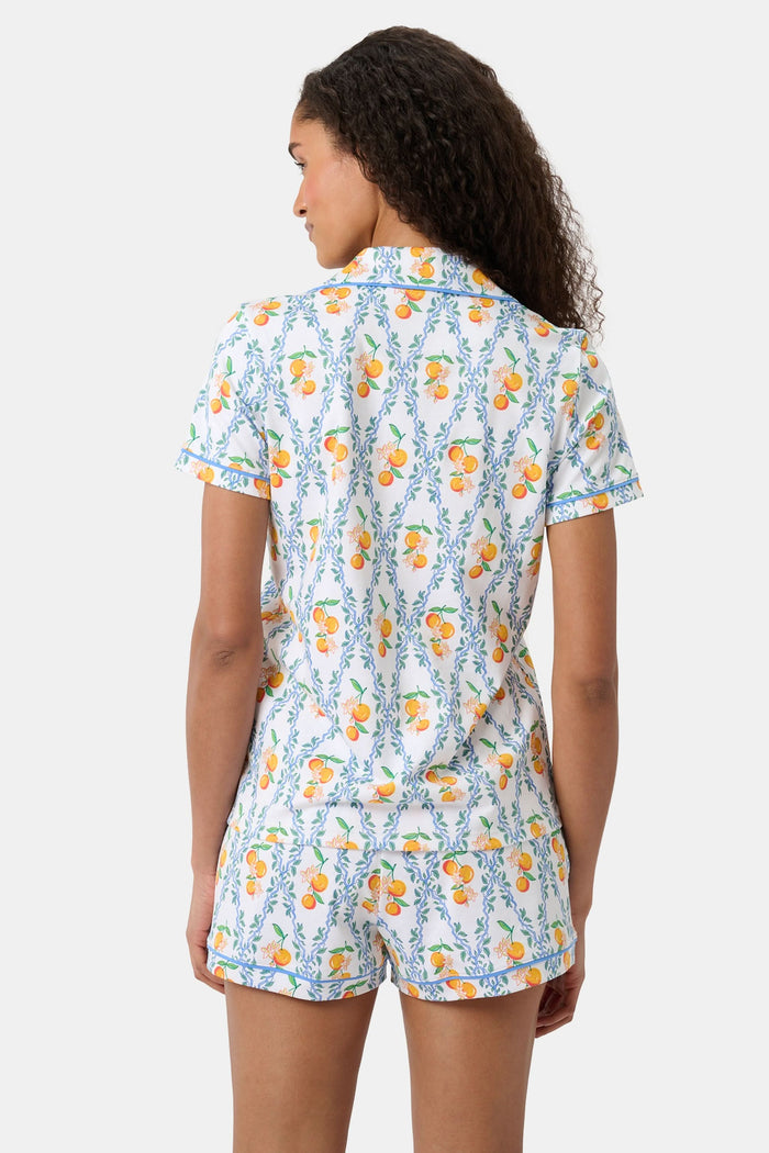 A woman with long hair strolls on the grass in a garden, wearing the Clementine Trellis Polo Pajamas by Roller Rabbit. Green plants and purple flowers bloom in the sunlight behind her.