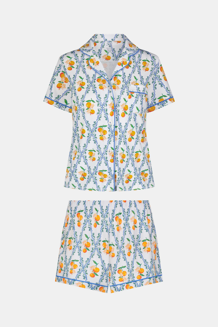 A woman with long hair strolls on the grass in a garden, wearing the Clementine Trellis Polo Pajamas by Roller Rabbit. Green plants and purple flowers bloom in the sunlight behind her.