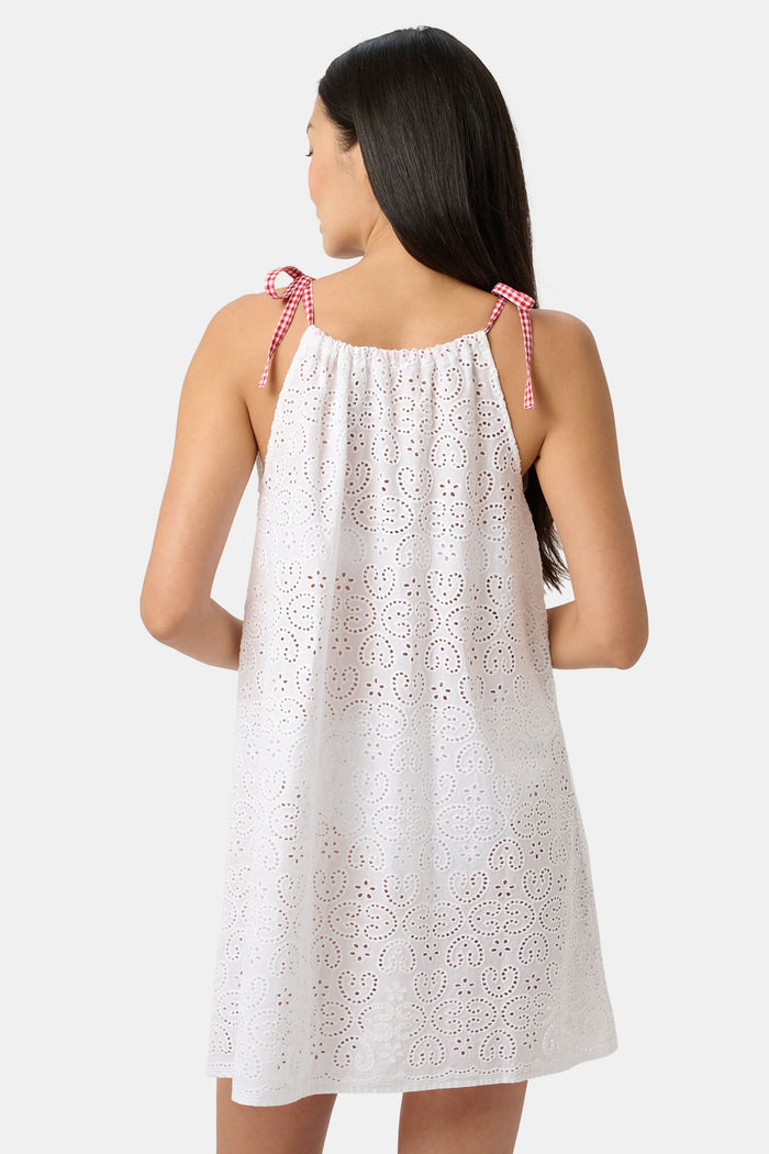A woman models the Roller Rabbit Clover Eyelet Caroline Cover Up, a white cotton dress with adjustable shoulder straps and red gingham ties, set against a plain light gray background.
