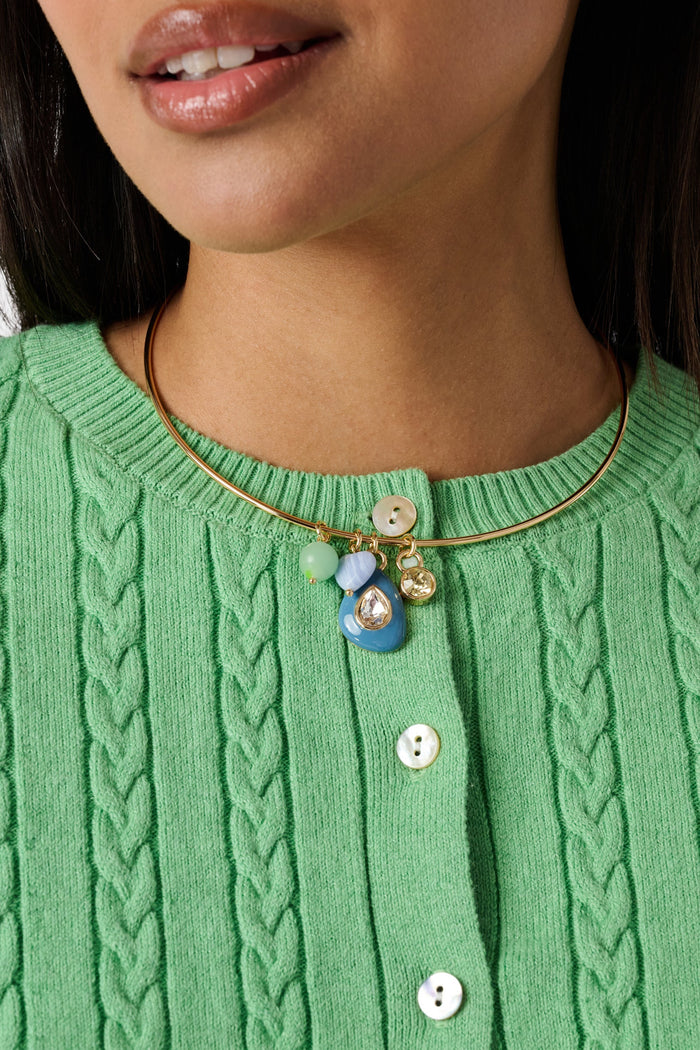A woman wears a green cable-knit sweater and the Roller Rabbit Collar Stone Necklace, which features multiple colorful charms, including a blue pendant and beads. Only her lower face, neck, and upper chest are visible.