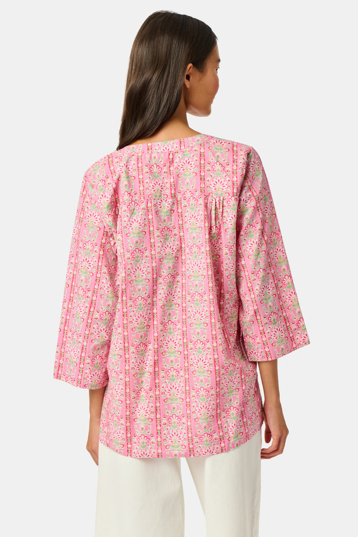 A woman wearing the Roller Rabbit Cora Floral Stripe Pippa Dress—a pink, short-sleeve, midi-length cotton dress with ruffled shoulders and a floral pattern—stands facing forward against a plain white background.