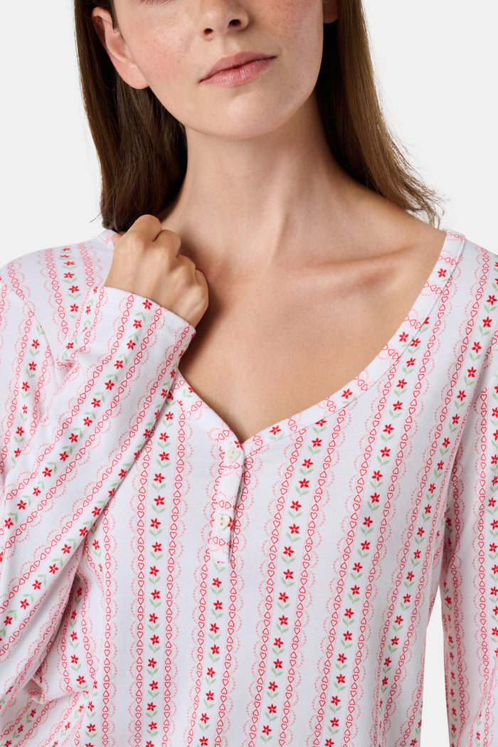 A woman smiles while wearing the Roller Rabbit Daisy Heart Stripe Jessie Long Sleeve Tee, a soft Pima cotton top adorned with pink and red daisies, paired with matching pajama bottoms, against a plain white background.