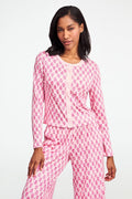 A woman stands against a white background, wearing the Daisy Paisley Nessa Cardigan by Roller Rabbit over a matching pink pajama set with red pineapple print, one hand on her hip.