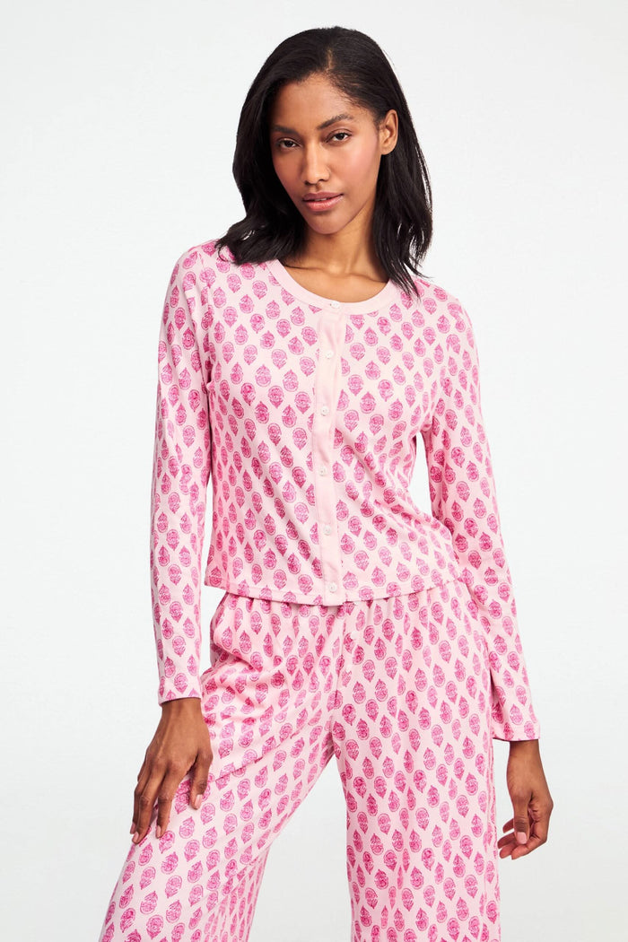 A woman stands against a white background, wearing the Daisy Paisley Nessa Cardigan by Roller Rabbit over a matching pink pajama set with red pineapple print, one hand on her hip.
