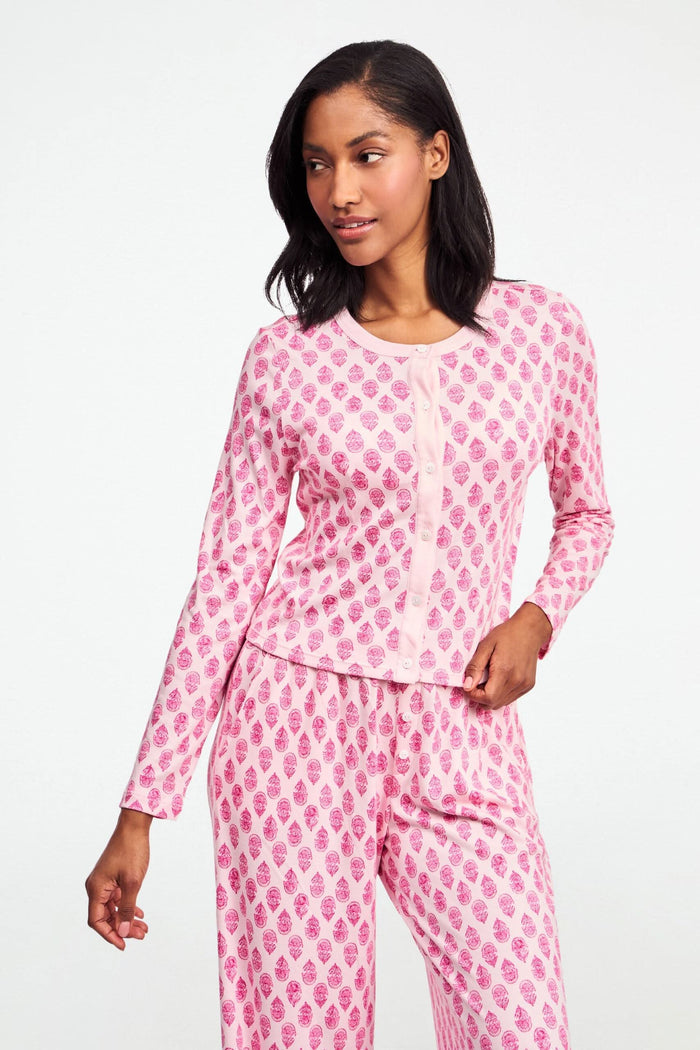 A woman stands against a white background, wearing the Daisy Paisley Nessa Cardigan by Roller Rabbit over a matching pink pajama set with red pineapple print, one hand on her hip.