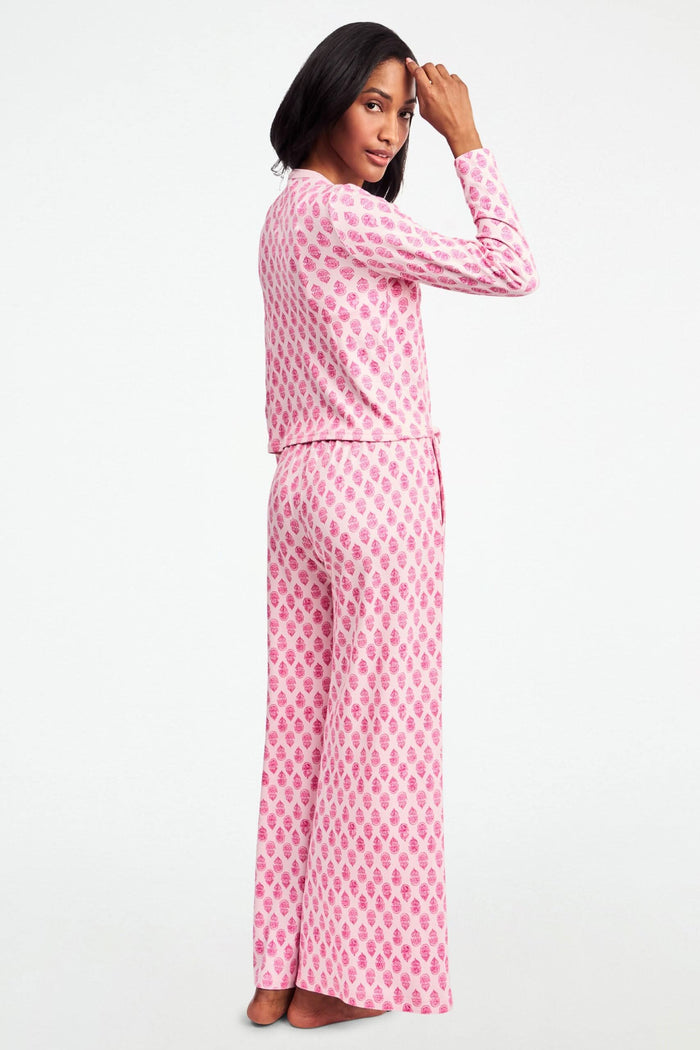 A woman stands against a white background, wearing the Daisy Paisley Nessa Cardigan by Roller Rabbit over a matching pink pajama set with red pineapple print, one hand on her hip.