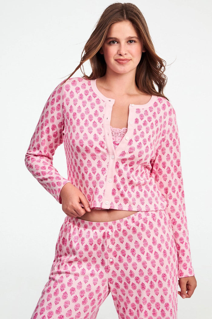 A woman stands against a white background, wearing the Daisy Paisley Nessa Cardigan by Roller Rabbit over a matching pink pajama set with red pineapple print, one hand on her hip.